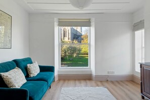 A room with a teal sofa, a white rug, and a window with a view of a building and greenery. - The Exmouth Nook, Walk to Beach, Parking Nearby (Exmouth)