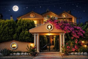 Ein zweistöckiges Haus mit überdachter Veranda, einem Mond am Himmel und einem Garten mit rosa Blumen.