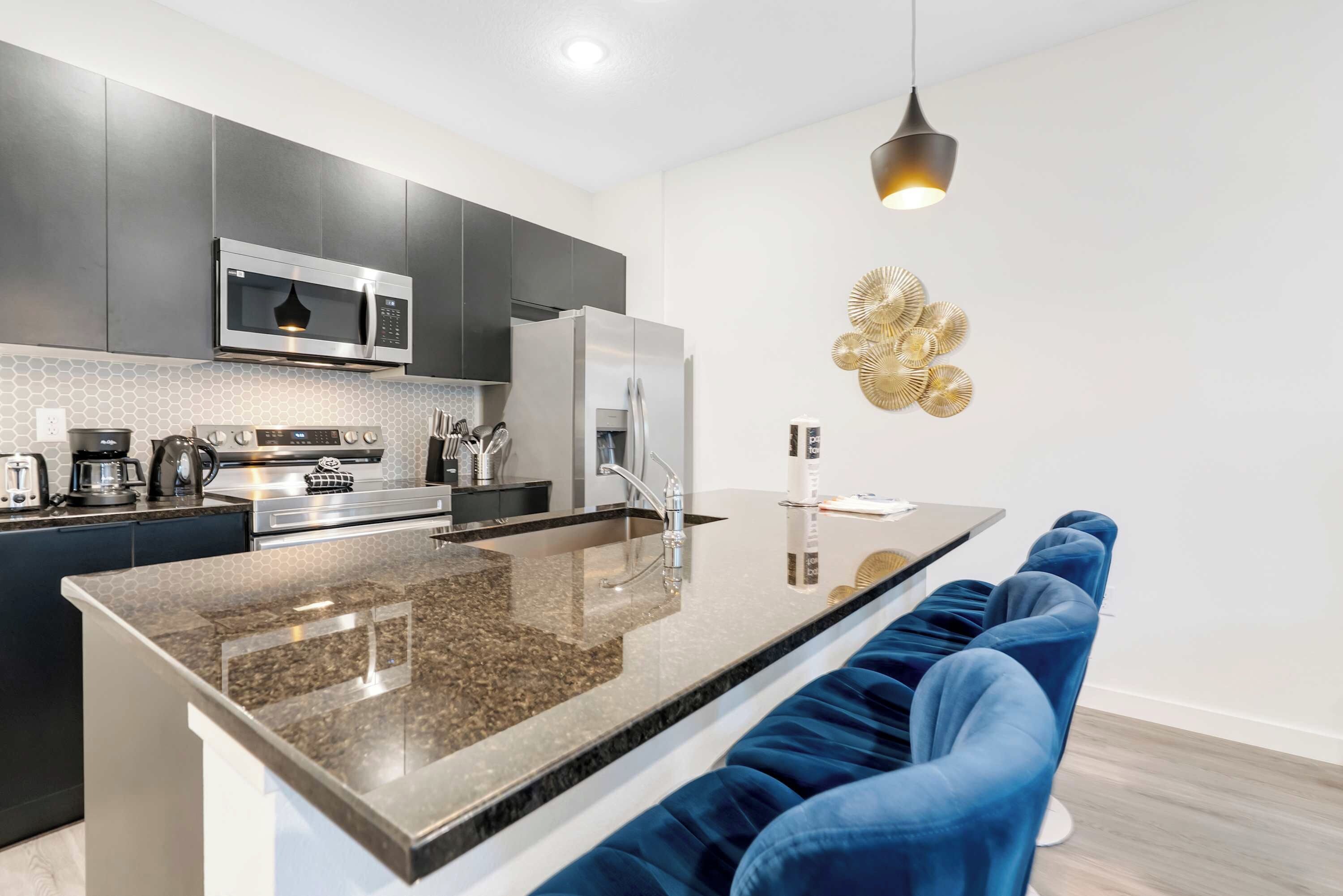 A modern kitchen with a central island, dark cabinetry, stainless steel appliances, and a seating area with blue upholstered chairs.