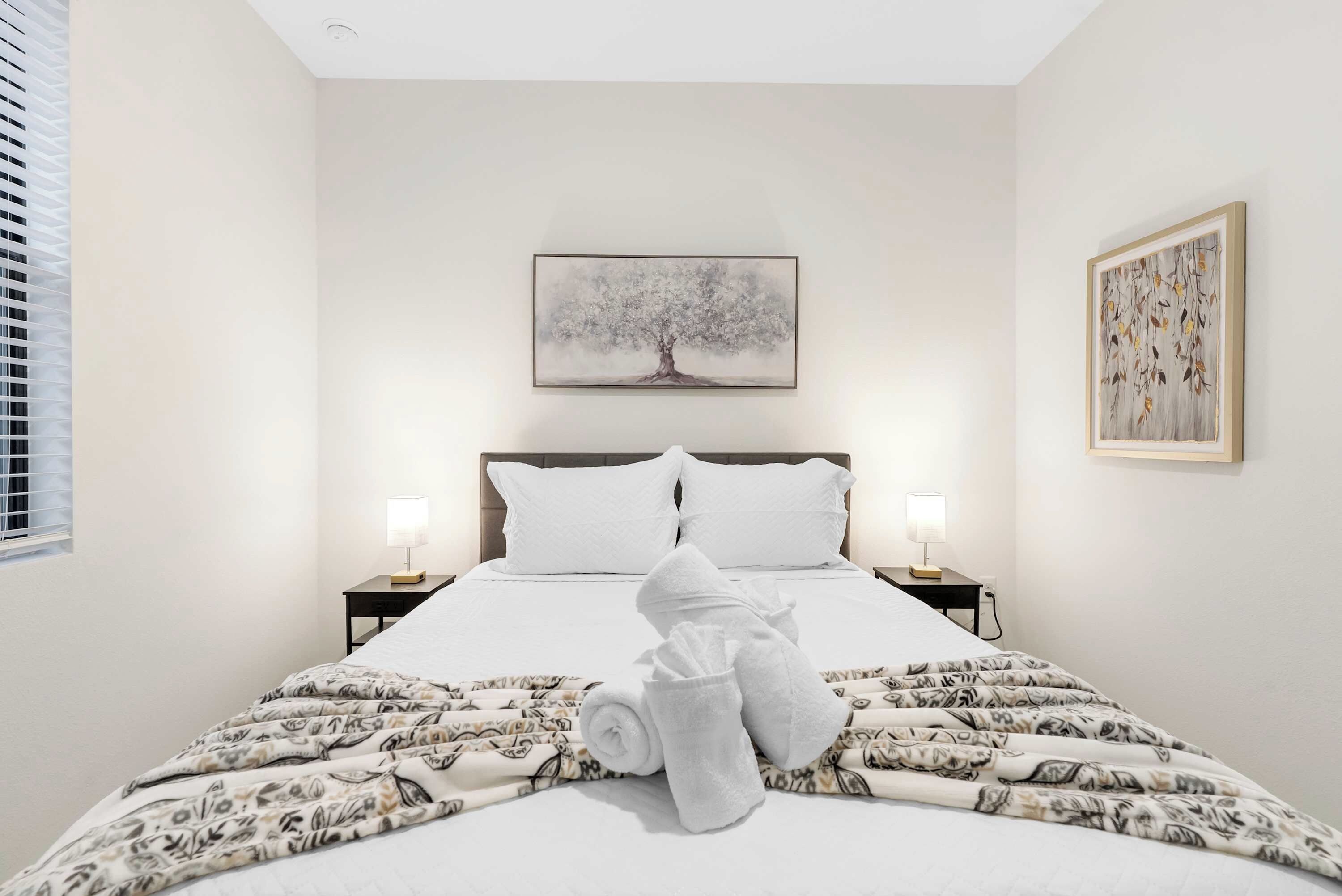 A hotel room with a bed, two bedside lamps, a painting above the bed, and a window with blinds.