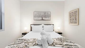 A hotel room with a bed, two bedside lamps, a painting above the bed, and a window with blinds.