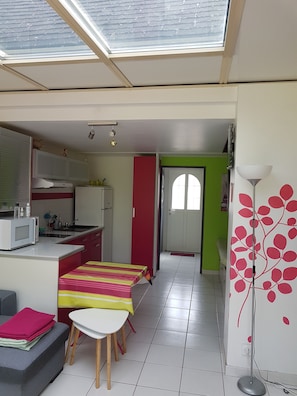 A compact kitchen with a microwave, sink, and cabinets. There is a dining area with a table and chairs, and a sofa. A red door leads to another room. - House in a complex with a swimming pool in the summer, 900 meters from the beach (Batz-sur-Mer)