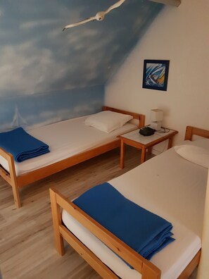 A room with two wooden beds, each with a blue blanket and white pillows. There is a nightstand with a lamp and a small table with a bag on it. The wall features a painted sky with clouds and a framed picture. - House in a complex with a swimming pool in the summer, 900 meters from the beach (Batz-sur-Mer)