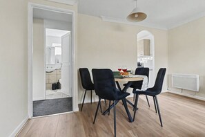 A dining area with a table set for two, four chairs, and a kitchen visible through an open door. - Comfortable Flat in Bedford – Sleeps 3–4 Guests (Bedford)