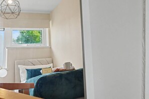 A bedroom with a bed, a window with a view of greenery, a bedside table with a cup, and a geometric light fixture. - Comfortable Flat in Bedford – Sleeps 3–4 Guests (Bedford)