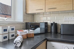 A kitchen with a microwave, toaster, and coffee maker. There are storage containers labeled with tea, milk, and sugar. A towel is draped over the counter. - Comfortable Flat in Bedford – Sleeps 3–4 Guests (Bedford)