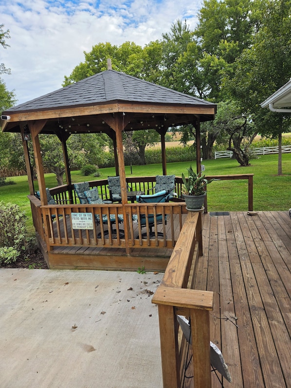 A wooden gazebo with a bench and a sign, surrounded by a deck and greenery.