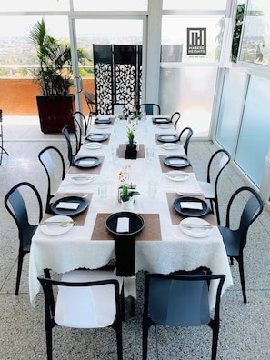 Un comedor con una larga mesa dispuesta para seis personas, rodeada de sillas, con una planta en maceta y paneles decorativos al fondo.