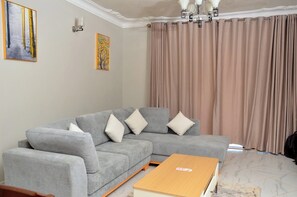 A living room with a grey sectional sofa, a wooden coffee table, and a large window with beige curtains.