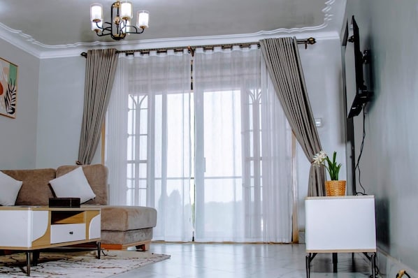 Una sala de estar con sofá, una mesita auxiliar, televisor de pantalla plana y un ventanal grande con cortinas.