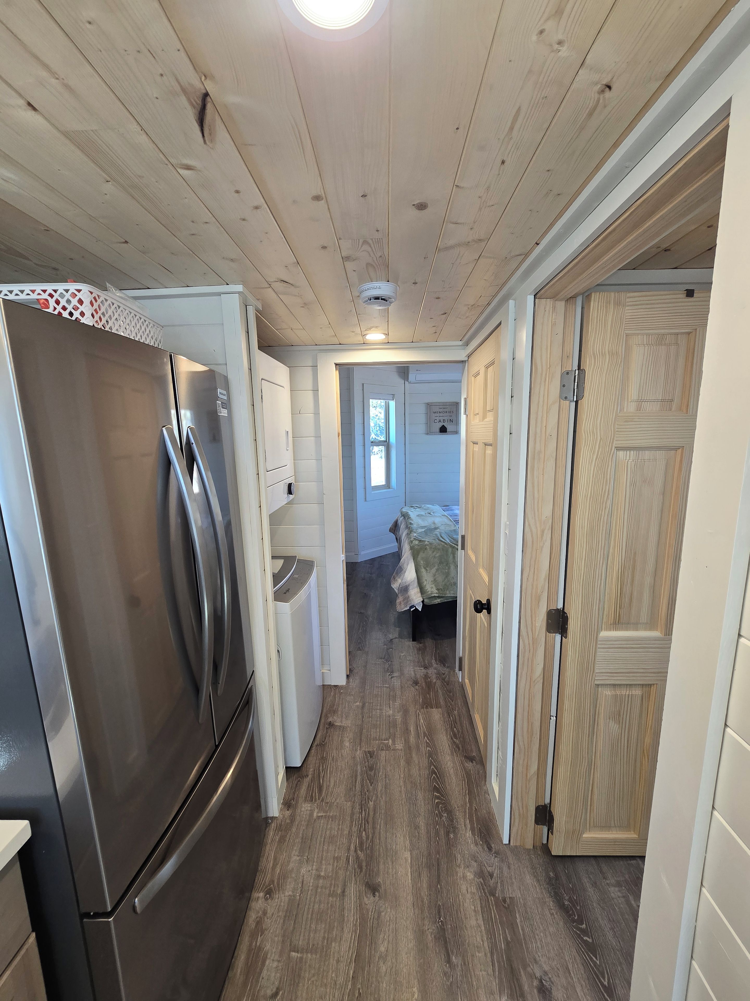 A narrow hallway with a stainless steel refrigerator on the left, wooden doors on the right, and a window at the end.