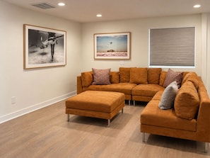 A living room with a sectional sofa, an ottoman, and two framed pictures on the wall.
