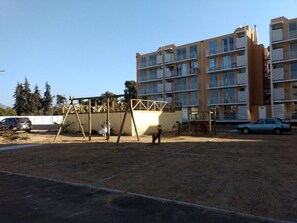 Un immeuble résidentiel doté de nombreux balcons, d’une aire de jeux avec des balançoires et d’un espace de stationnement.