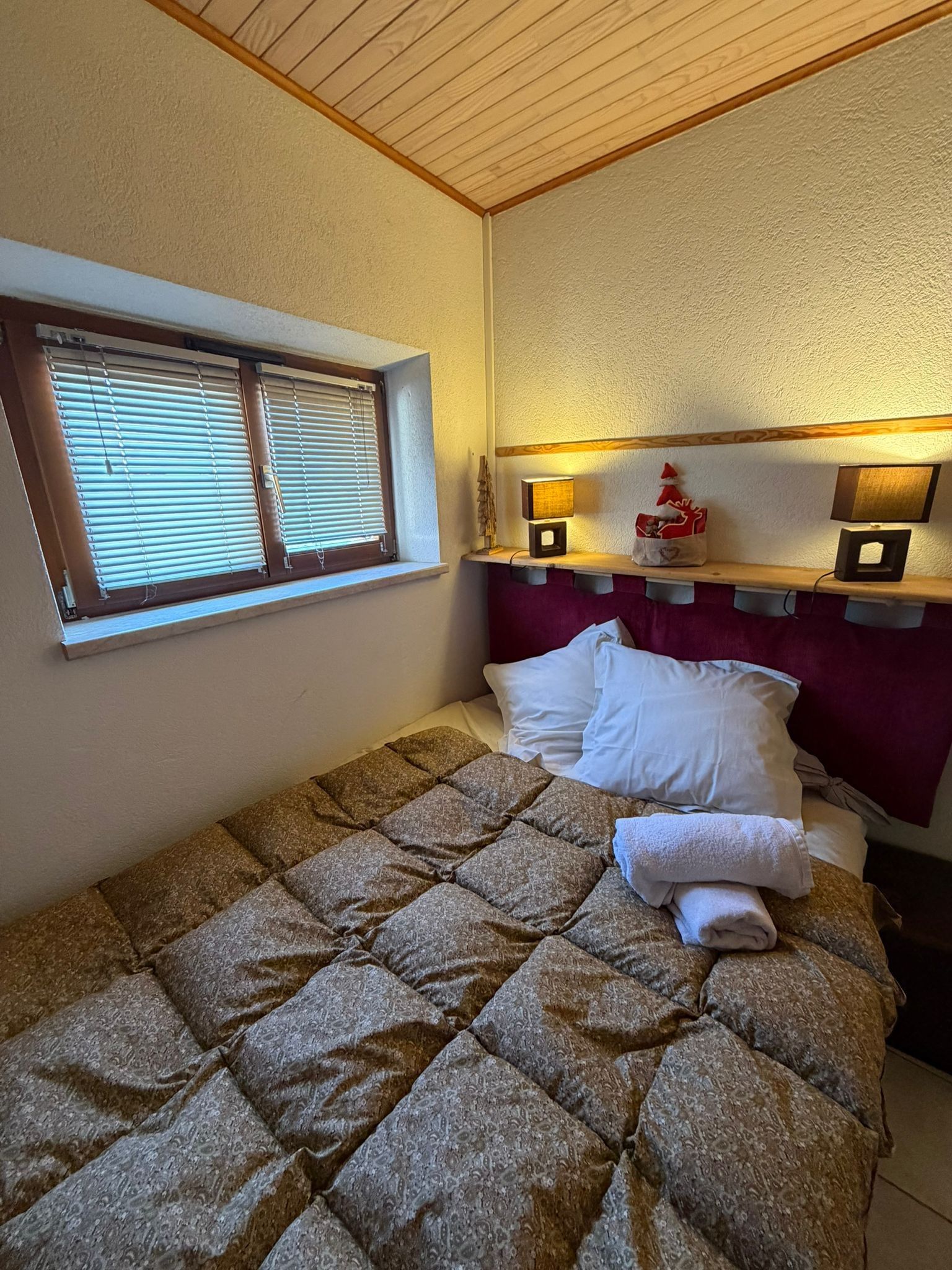 A single bed with a quilted cover, a window with blinds, and two wall-mounted lamps on a wooden shelf above the bed.
