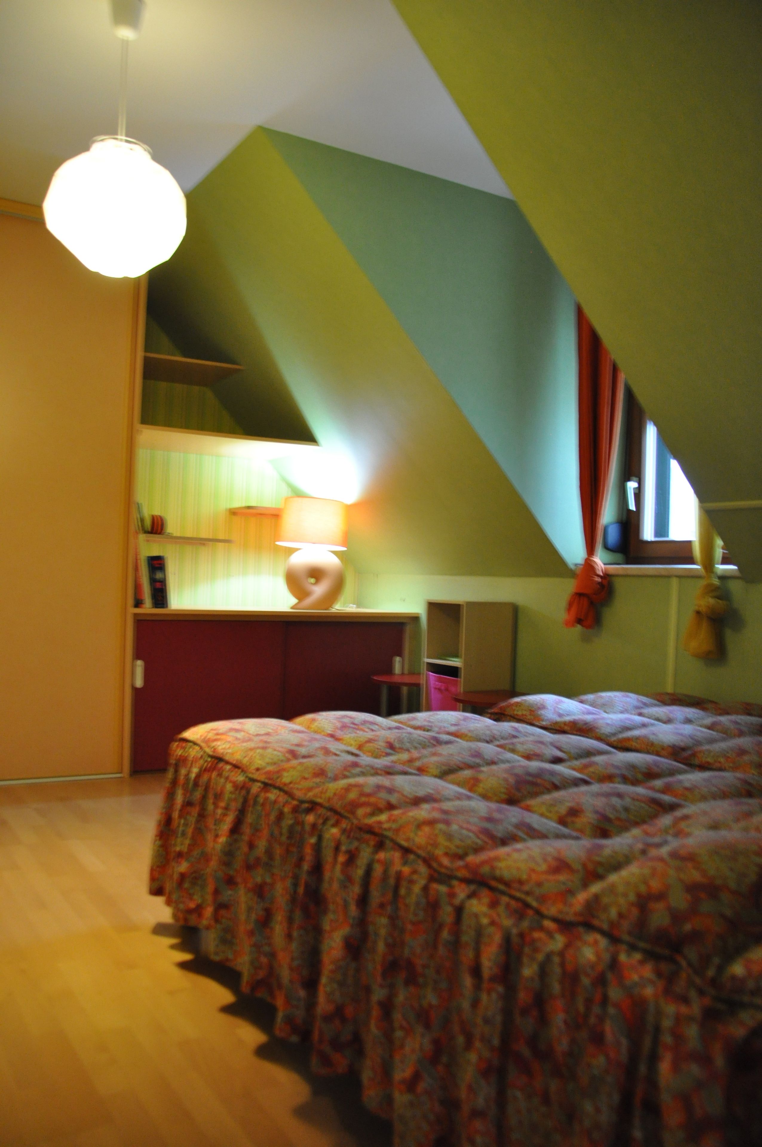 Ett sovrum med ett sluttande tak, en säng med ett mönstrat överkast, ett skrivbord med en lampa och ett fönster med gardiner.