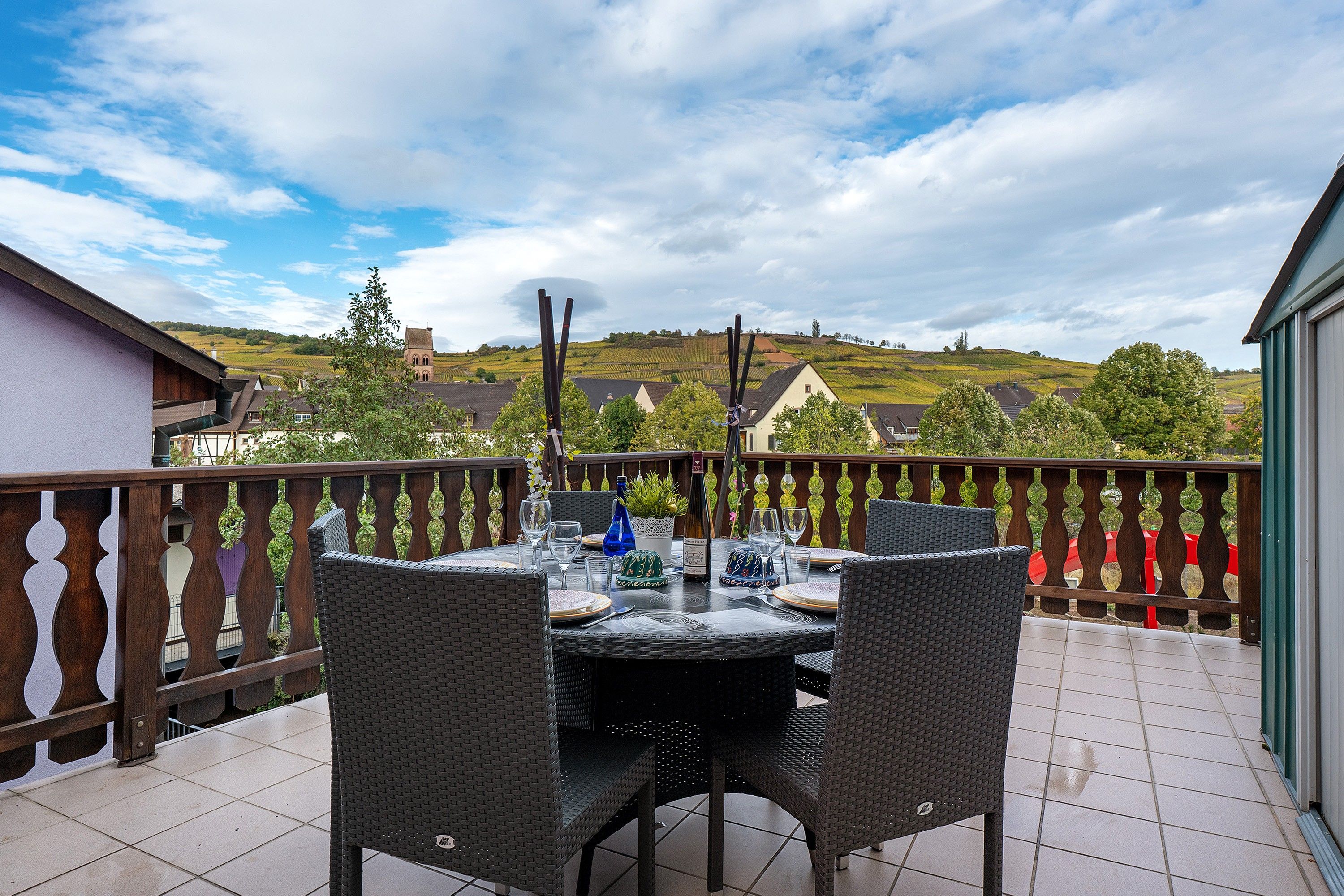 En terrasse med et spisebord omgivet af fletstole, med udsigt over et naturskønt landskab med huse og bakker.