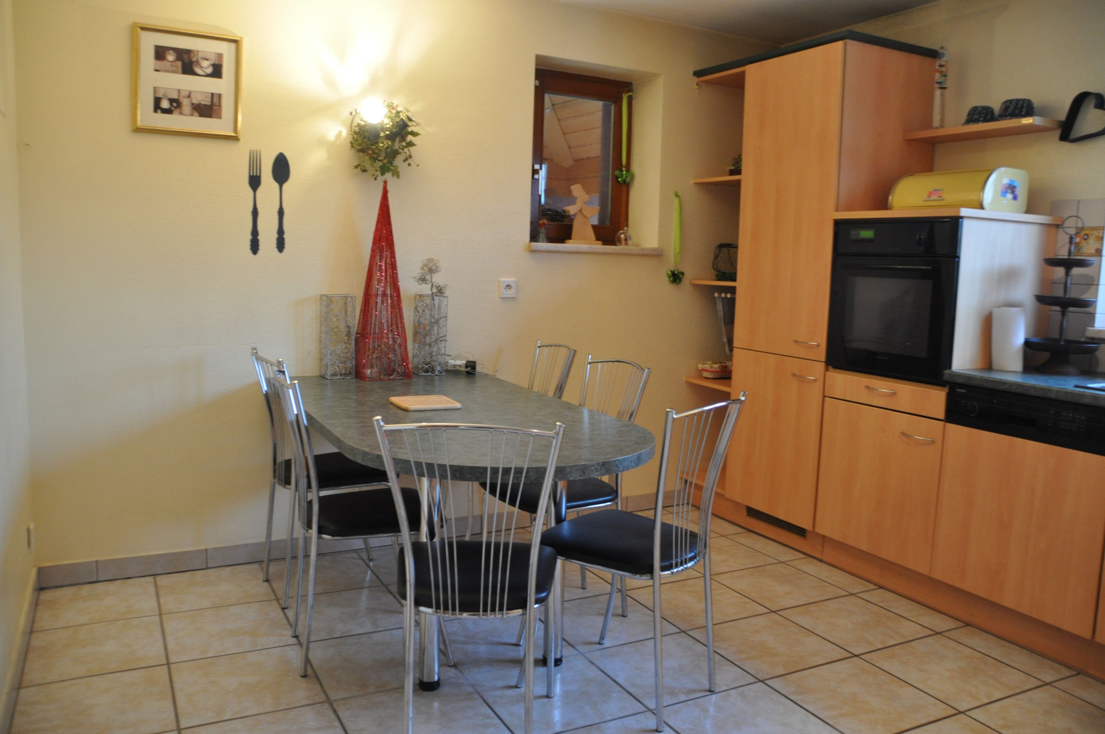 Un comedor con mesa redonda y sillas, una cocina con alacenas de madera y electrodomésticos, y un aplique de pared.