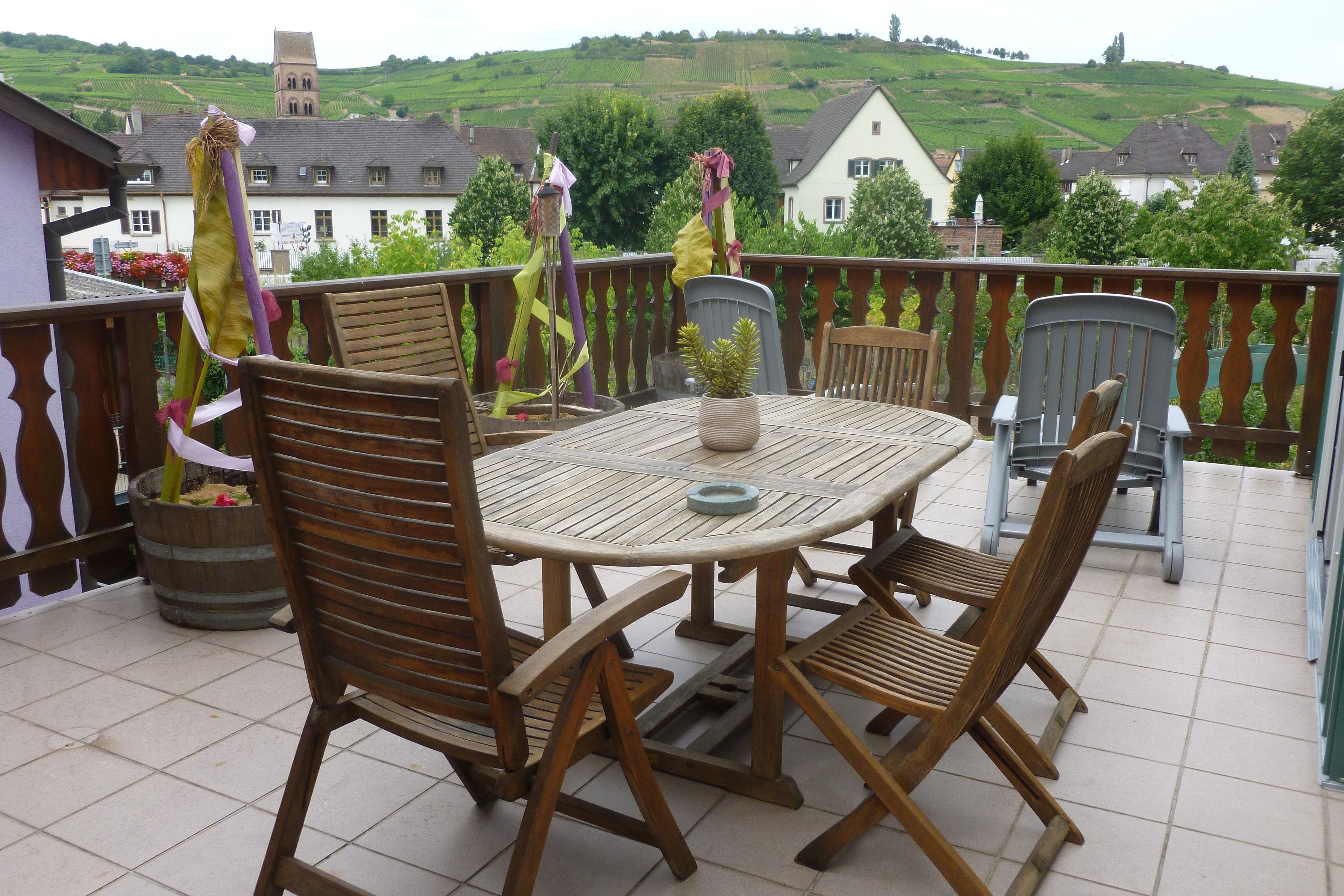 A wooden deck with a table and chairs, overlooking a vineyard and houses.