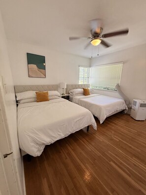 Een kamer met twee bedden, een plafondventilator, een raam met zonwering en een wanddecoratie.