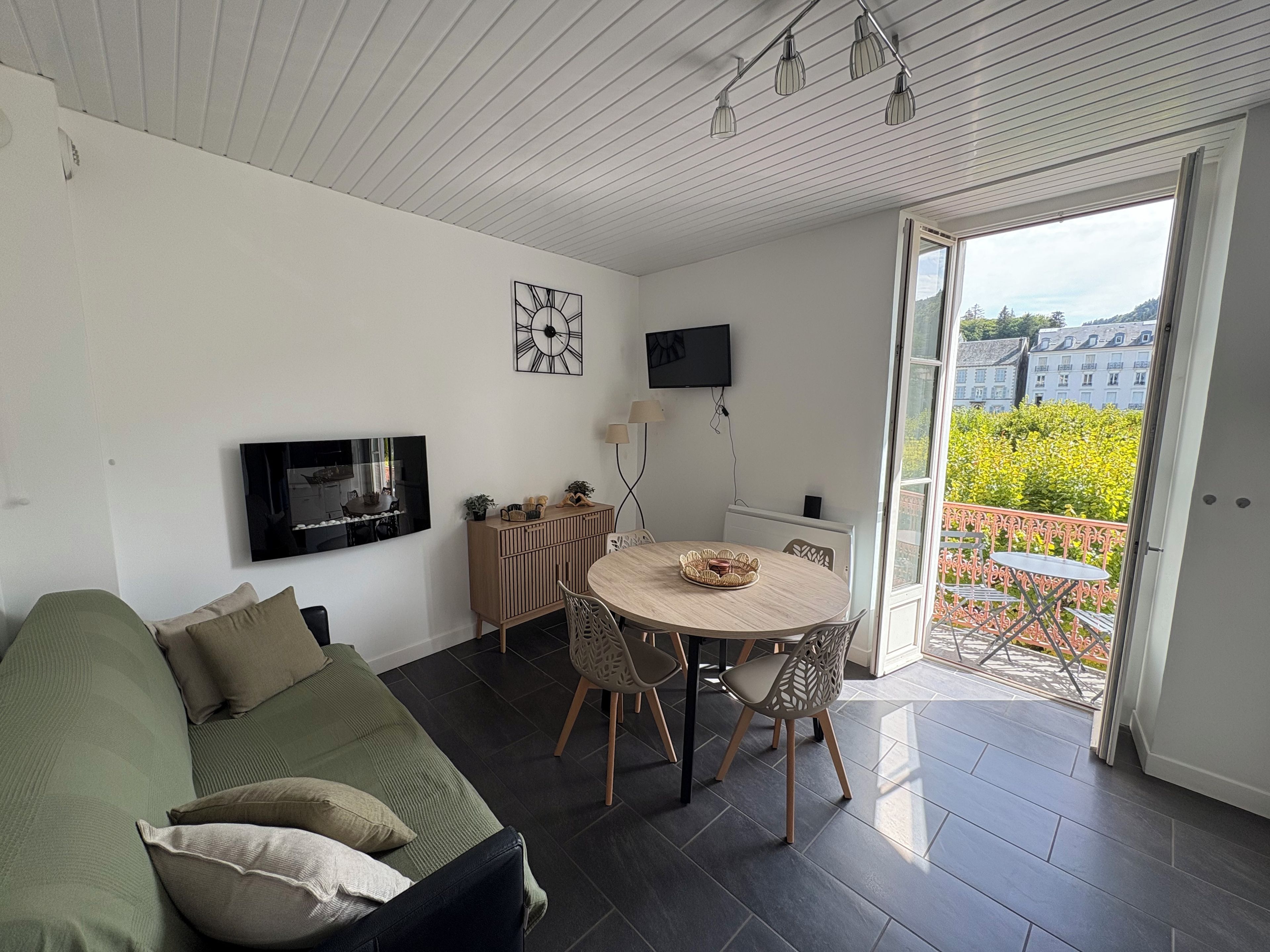 Un salon moderne comprenant un canapé, une table à manger avec des chaises, un téléviseur à écran plat fixé au mur et un balcon offrant une vue sur les bâtiments et la verdure.