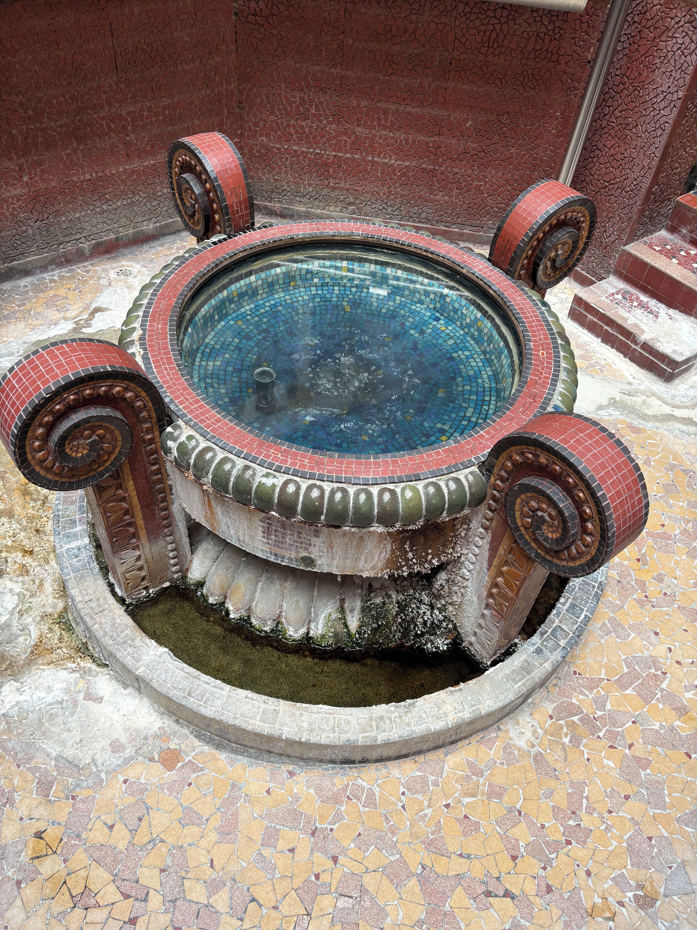 A circular hot tub with four legs, surrounded by a tiled floor with a mosaic pattern.
