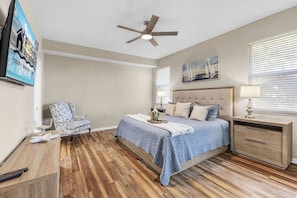 Une chambre à coucher comprenant un lit, une chaise, une table de chevet, une lampe, un ventilateur de plafond et un tableau accroché au mur.