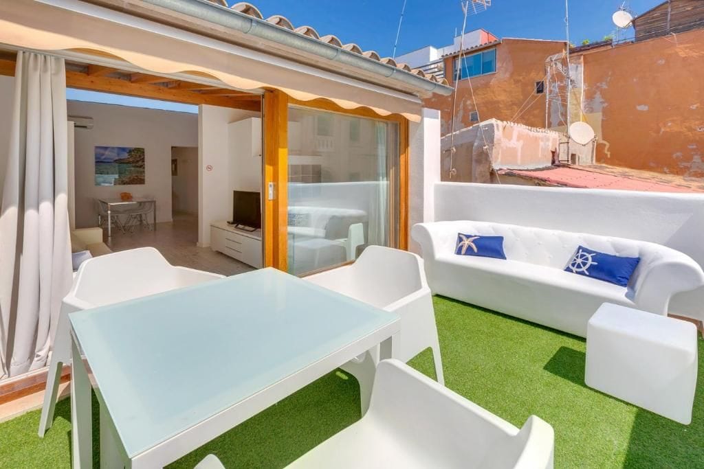 Un patio con una mesa de vidrio y sillas blancas, un sofá blanco con cojines azules, y vista a un edificio en el fondo.