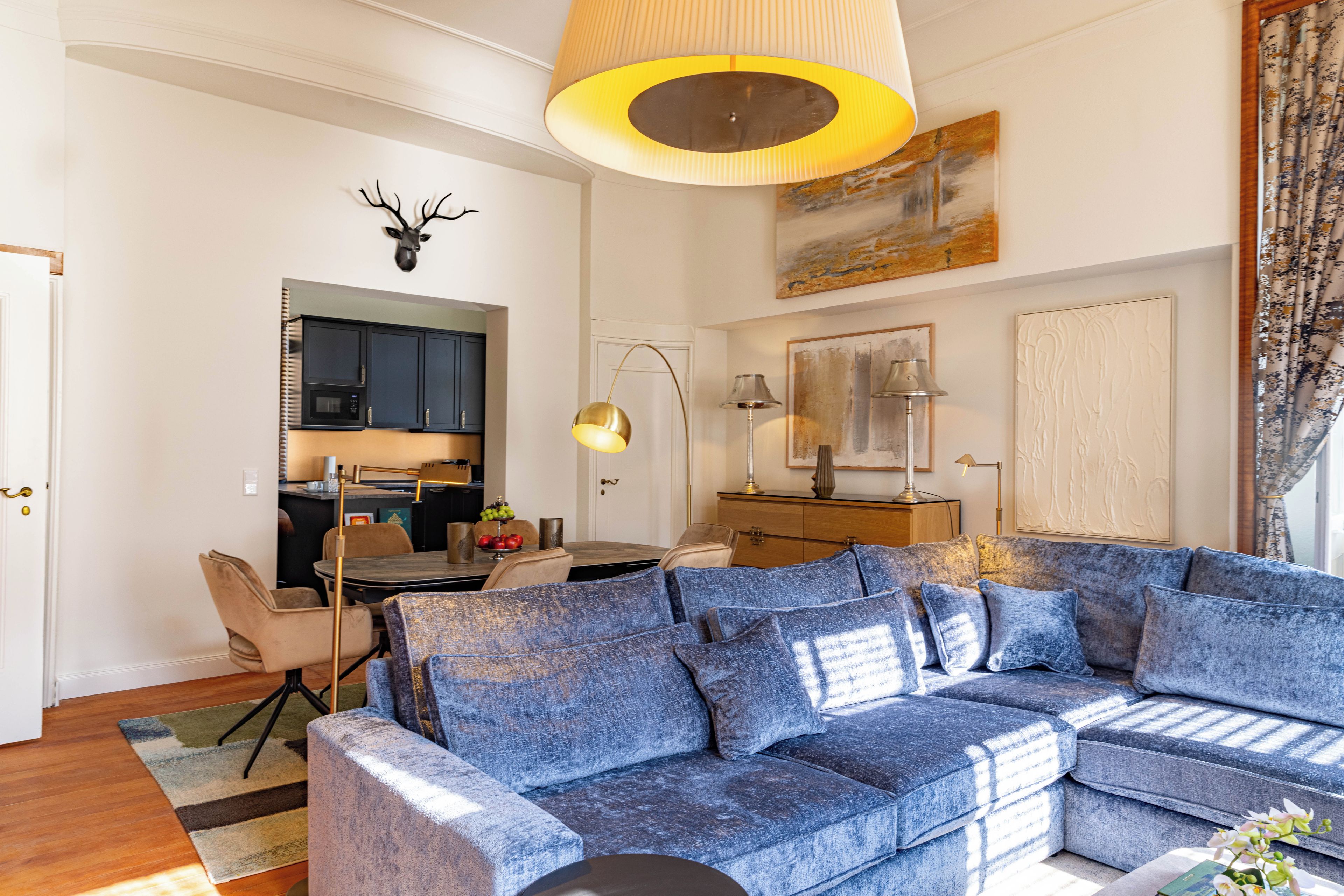 Ein Wohnzimmer mit einem blauen Sofa, einem Esstisch mit Stühlen, einer Küchenzeile und einem aufgehängten Hirschkopf an der Wand.
