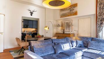 Ein Wohnzimmer mit einem blauen Sofa, einem Esstisch mit Stühlen, einer Küchenzeile und einem aufgehängten Hirschkopf an der Wand.