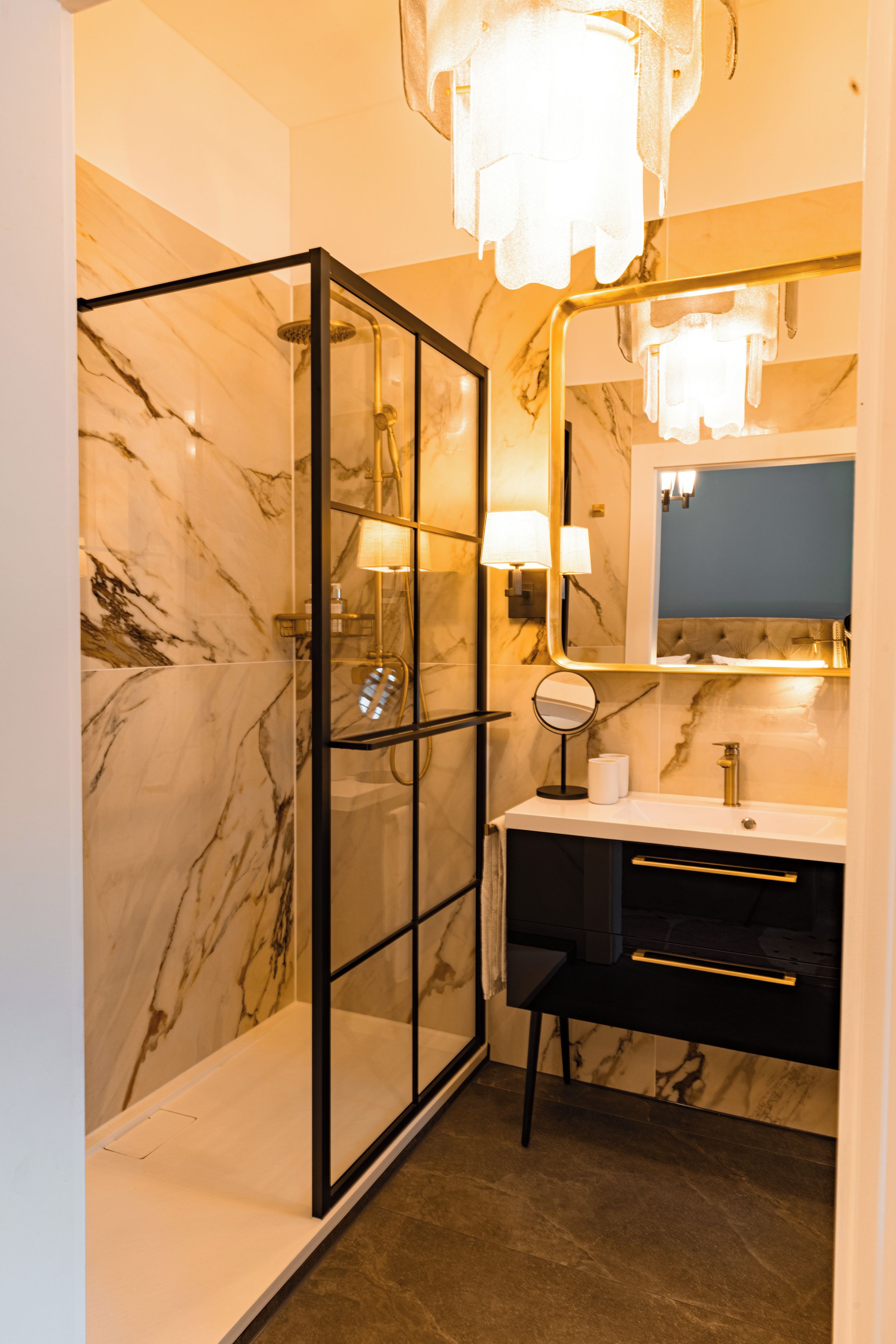 A modern bathroom with a glass-enclosed shower, marble walls, a vanity with a mirror, and a chandelier.