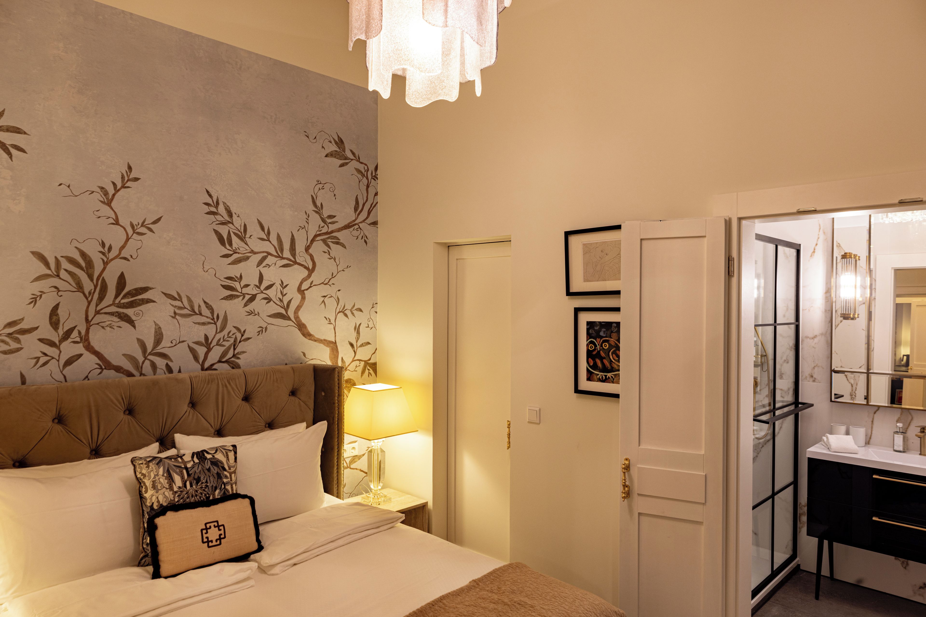 Habitación de hotel con cama, mesita de noche, lámpara, pared con diseño de árbol, puerta y baño visible a través de un panel de vidrio.