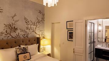 Habitación de hotel con cama, mesita de noche, lámpara, pared con diseño de árbol, puerta y baño visible a través de un panel de vidrio.