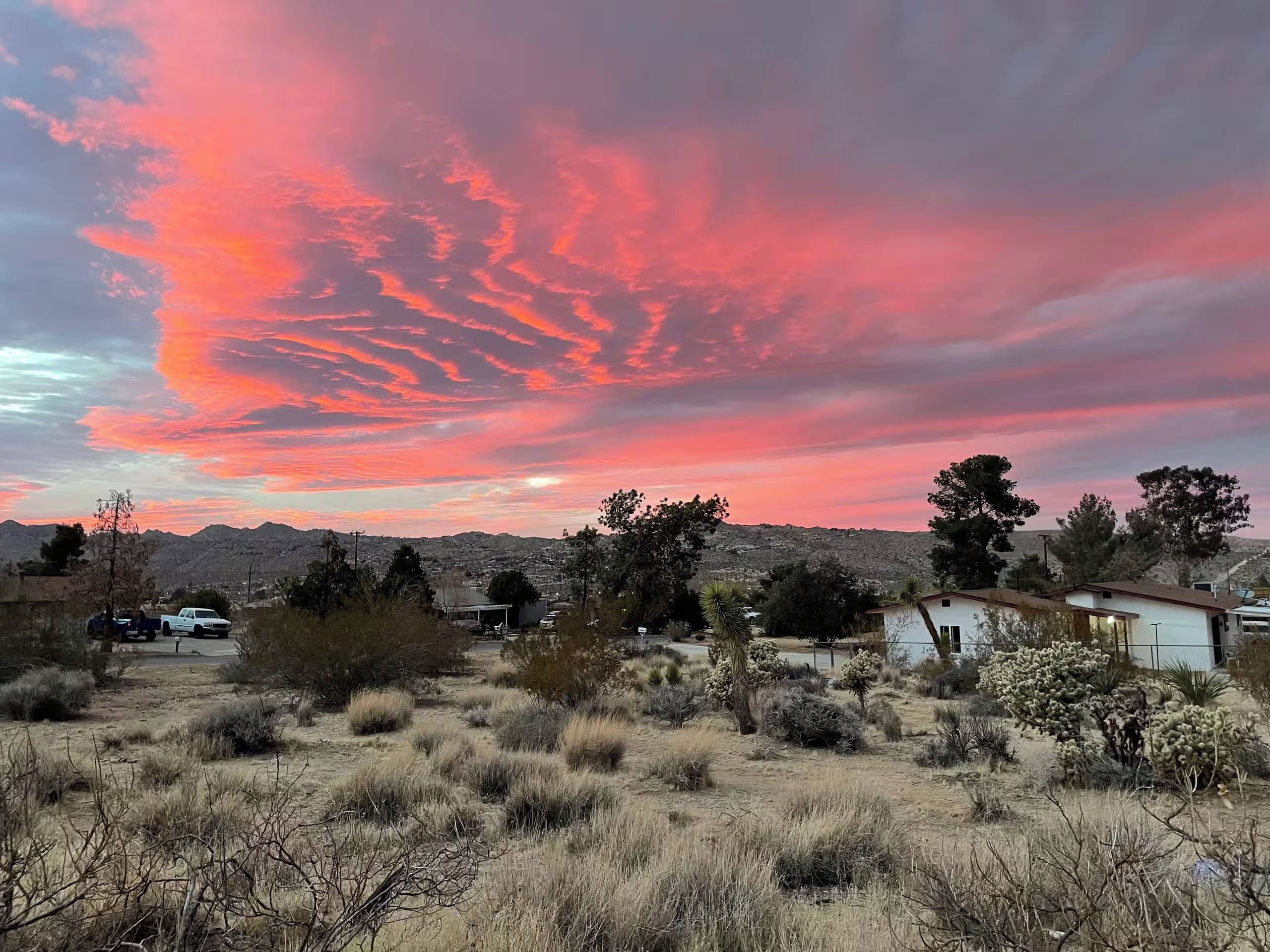 Un paysage désertique avec une végétation clairsemée, un bâtiment blanc et, en arrière-plan, des montagnes se dressant sous un ciel coloré au coucher du soleil.
