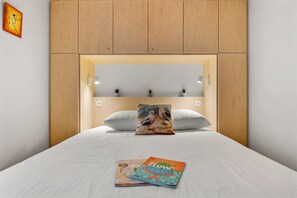Hotellihuone, jossa on sänky, tyynyjä, lehti ja seinään kiinnitetty valaisin.
