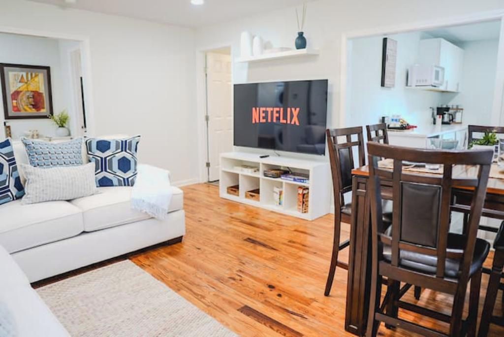 Una sala de estar moderna con un sofá blanco, cojines estampados, un televisor de pantalla plana, una mesa de comedor de madera con sillas y una zona de cocina al fondo.