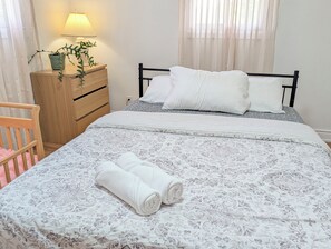 A neatly made bed with a floral-patterned duvet, a bedside lamp, a wooden nightstand with a potted plant, and a wooden chair with a pink cushion. - The Quinte House - Group Friendly with Pool Table (Quinte West)