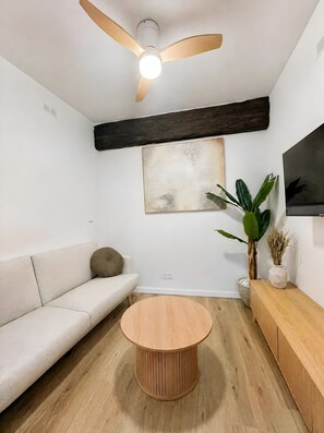 Un salon moderne avec un ventilateur de plafond, une table basse ronde en bois, un canapé blanc, un meuble en bois, une grande peinture abstraite et une plante en pot.