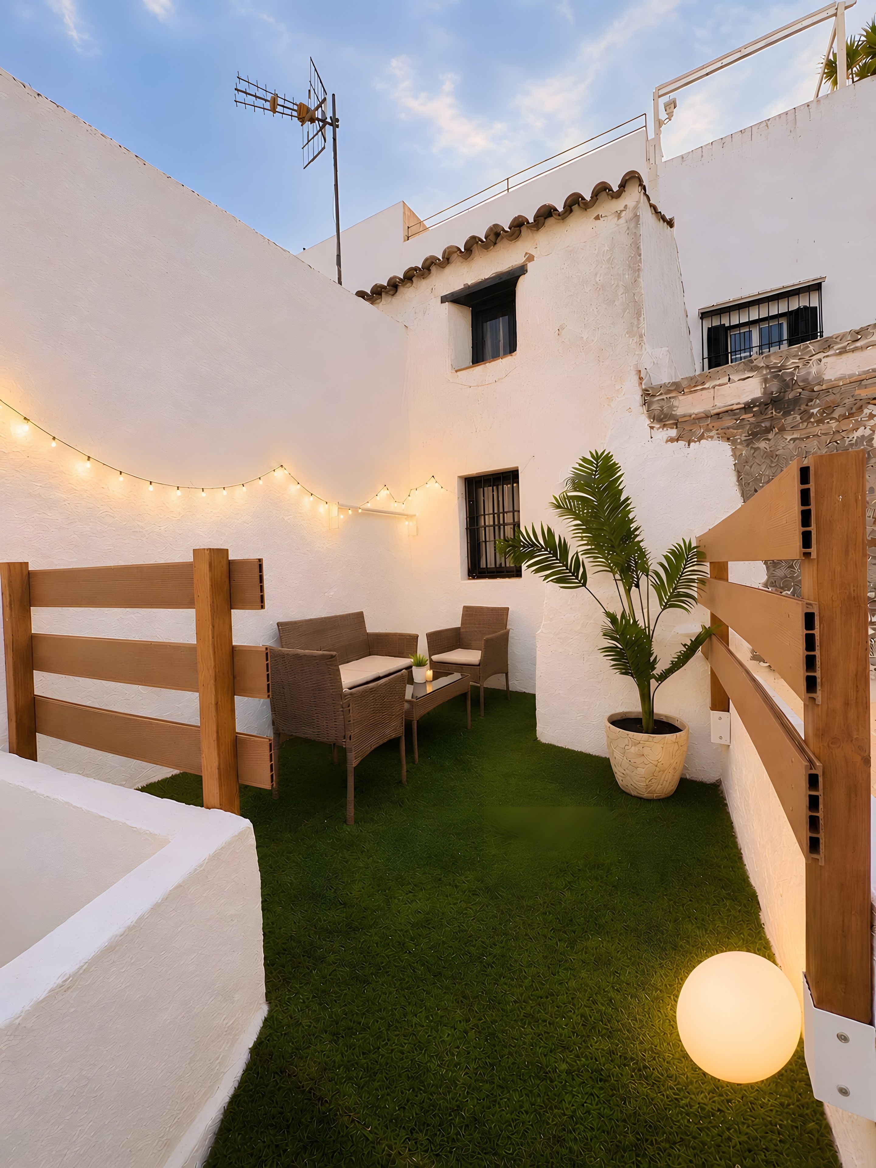 Eine Terrasse mit weißer Wand, einem kleinen Tisch, zwei Korbstühlen, einem Topfpflanzen und Lichterketten.