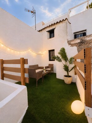 Une terrasse avec un mur blanc, une petite table, deux chaises en osier, une plante en pot et des guirlandes lumineuses.