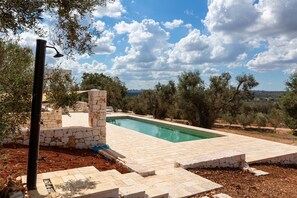 Une piscine avec des marches et un mur en pierre, entourée d’arbres et sous un ciel dégagé.