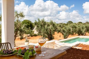 A table with a glass of wine, a plate of grapes, and a bowl of cherries, set against a backdrop of an olive grove and a swimming pool. - Casa tra gli Ulivi by Perle di Puglia (Ostuni)