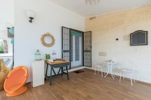 A room with a wooden floor, a dining table with chairs, a small white table with a coffee pot, and a large window with a view of greenery. - Villa Carolyta by BarbarHouse (Francavilla Fontana)