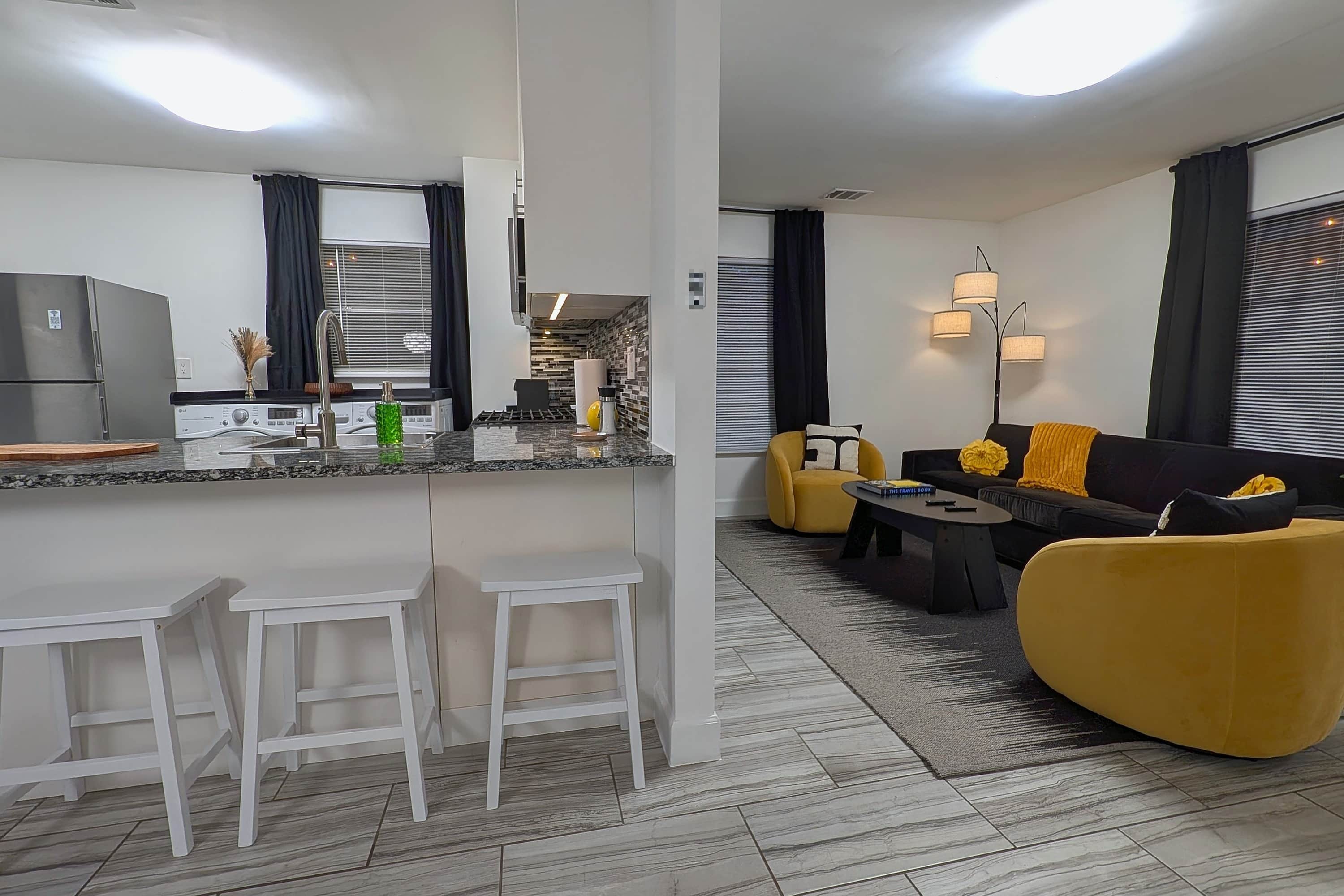 Een moderne woonkamer met een bank, gele fauteuils, een salontafel en een eethoek, met een keuken op de achtergrond.