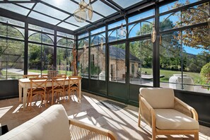 A conservatory with a table and chairs, large windows, and a view of a garden and building. - Château 'Le Chateau Des Girards' authentically renovated (Lans-en-Vercors)