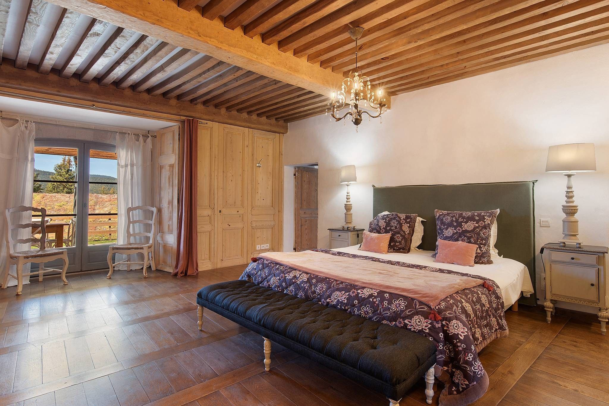 A bedroom with a wooden ceiling, a bed with floral bedding, a bench, a nightstand, and a view through a door.