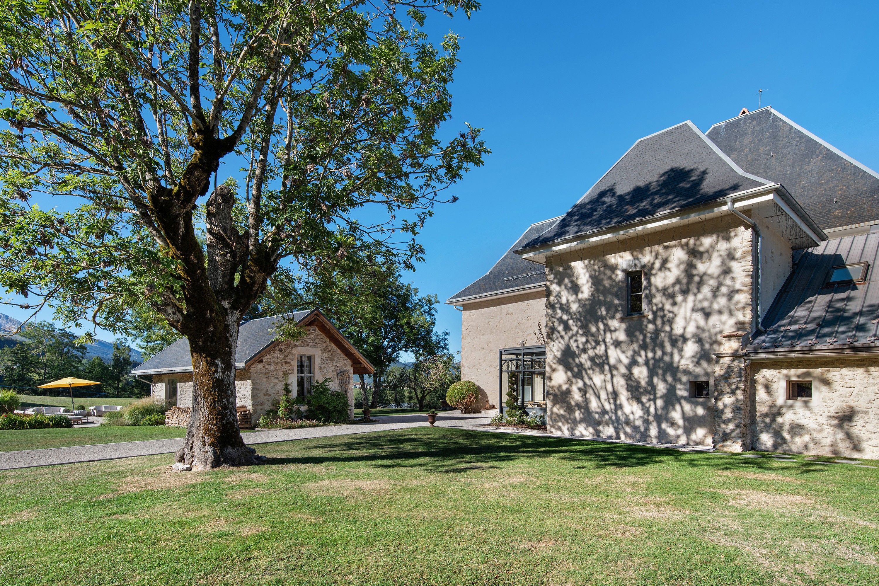 A stone building with a sloped roof, a large tree in the foreground, and a clear sky.