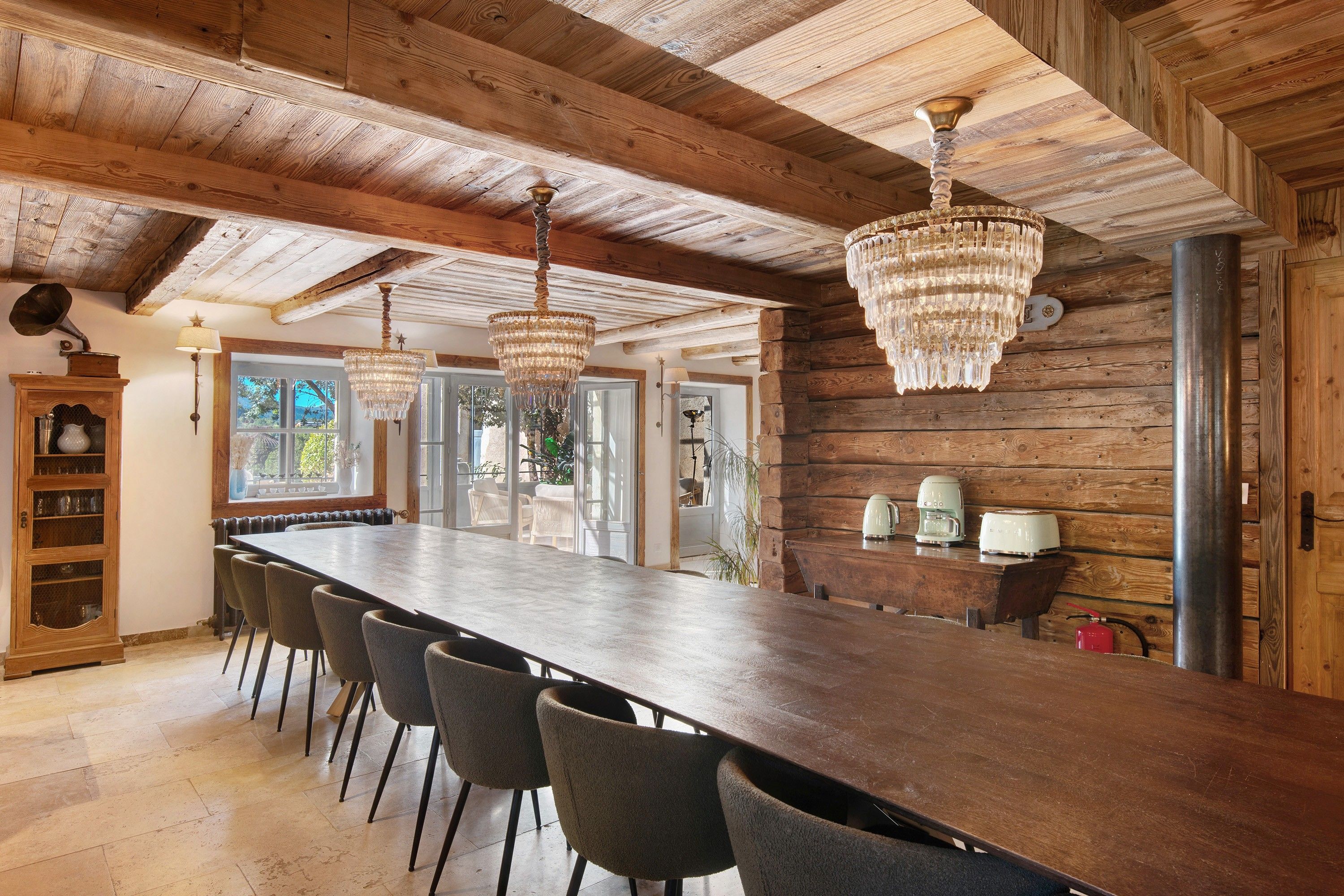 Une longue table à manger en bois, avec des chaises, un lustre et un gramophone d’époque.