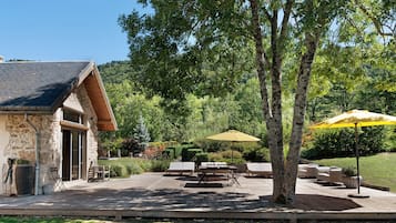 Un bâtiment en pierre doté d’un toit en pente, d’un espace extérieur aménagé avec des tables et des chaises, et d’un grand parasol offrant de l’ombre.