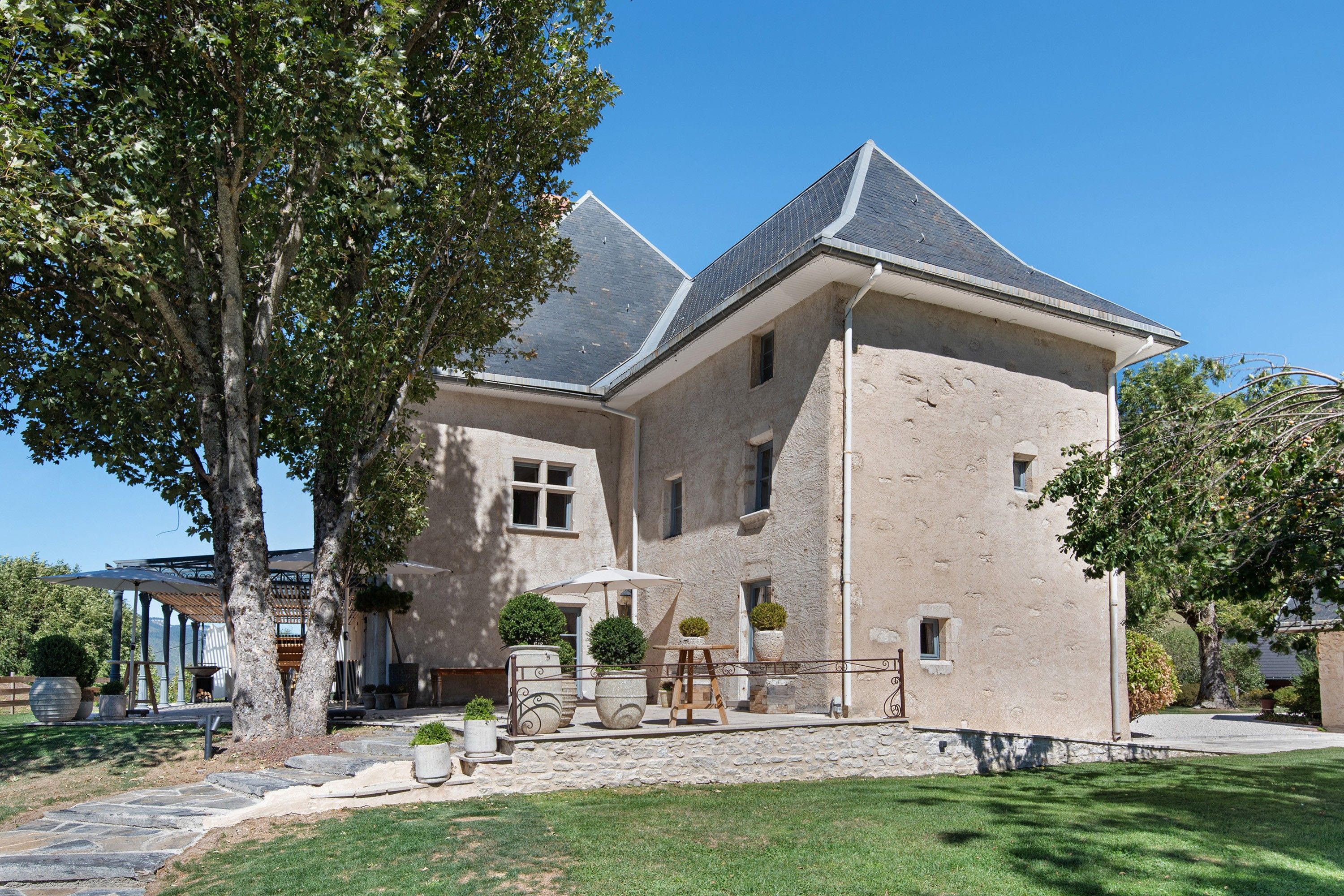 Une grande maison à deux étages, dotée d’une façade en pierre, d’un toit en pente et d’un jardin bien entretenu avec un espace patio.