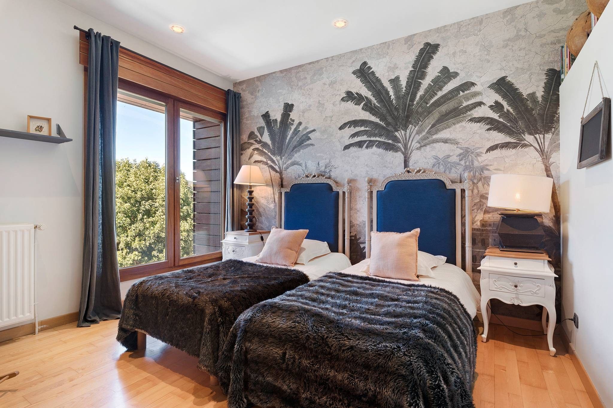 A bedroom with two beds, a large window, and a mural of palm trees.